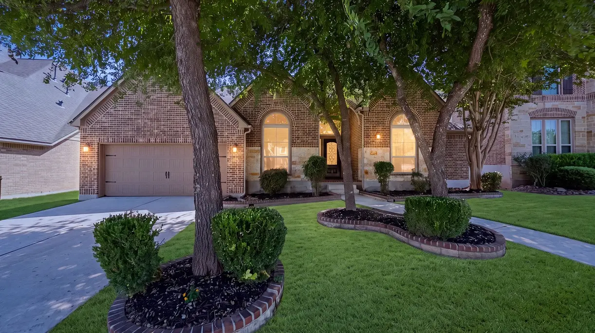 Front exterior of 13502 Palatine Hl, San Antonio, TX, featuring a brick-and-stone façade, mature shade trees, manicured landscaping, and a two-car garage at this 4-bedroom Perry Home in Bella Vista.
